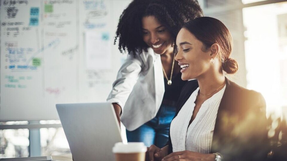 Twee vrouwen kijken lachend naar een laptop, met een whiteboard op de achtergrond.