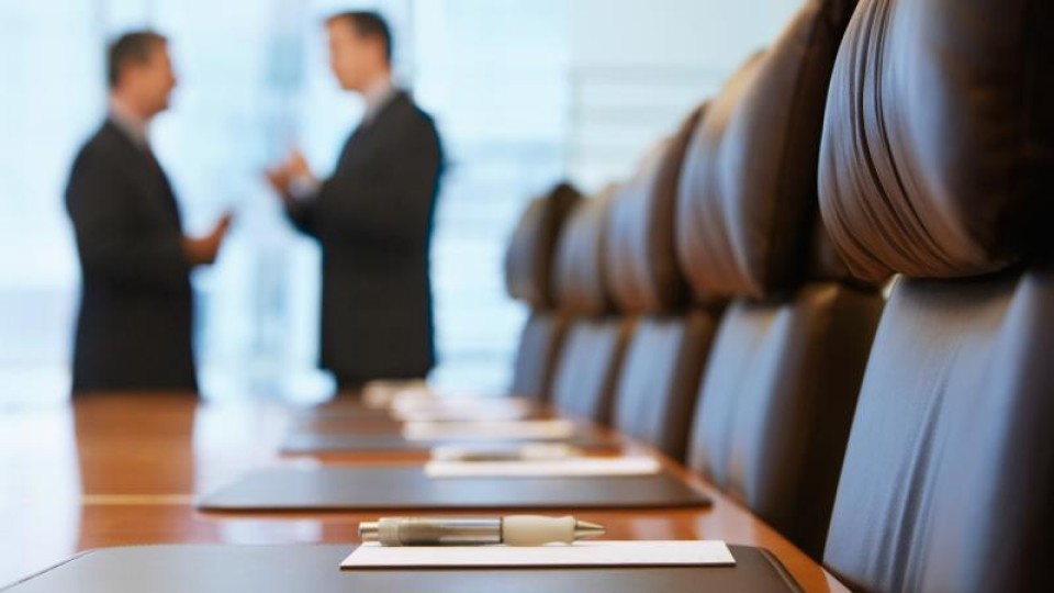 Een legen bestuurstafel met aan het raam twee mannen in discussie