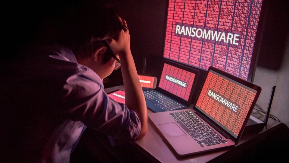 Persoon met hand op hoofd kijkt naar laptops en schermen met rode ransomware-tekst en binaire code.