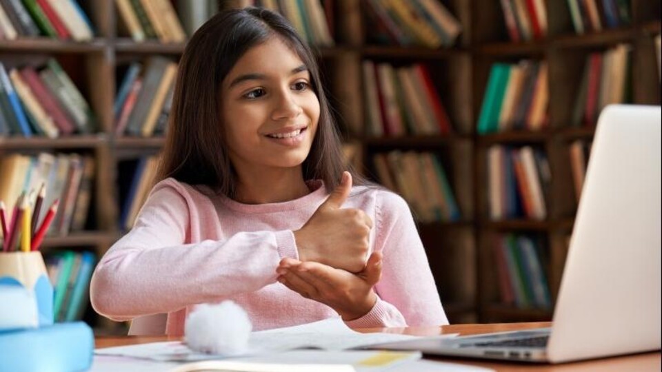 Jong meisje in roze trui maakt duim omhoog gebaar voor laptop met boeken op achtergrond