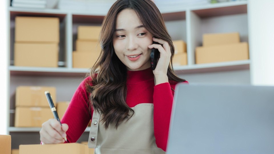 Vrouw noteert bestelling met pen met een telefoon tegen het oor en terwijl ze kijkt naar een laptopscherm