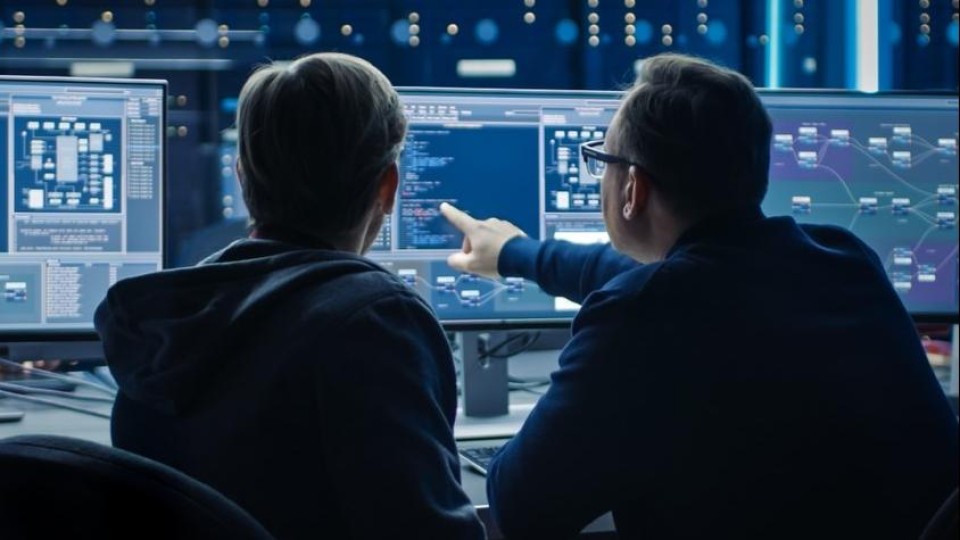 Twee mannen kijken naar een reeks beeldschermen met code en stroomdiagrammen