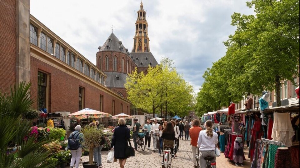 Mensen wandelen en fietsen op een marktstraat met marktkramen en een kerk met toren in de achtergrond.