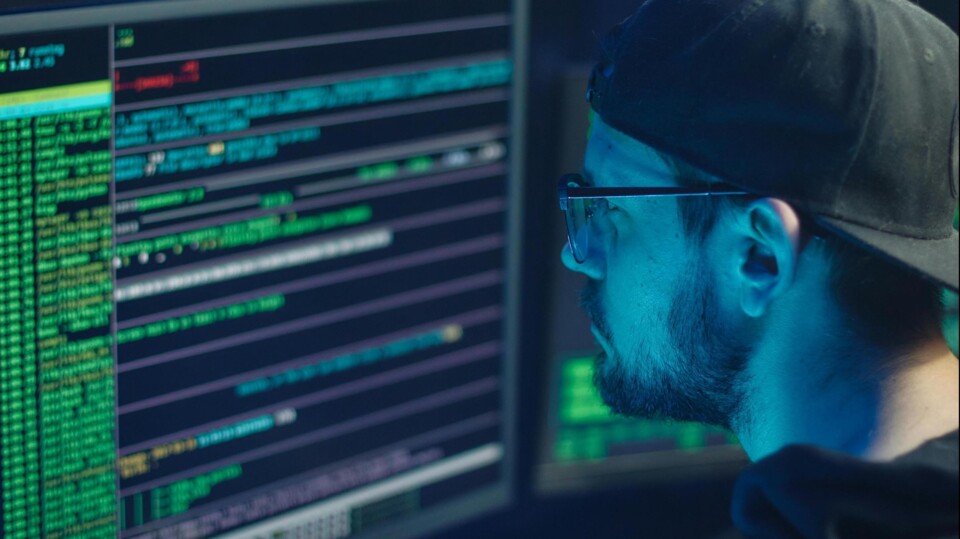 Man met pet en bril kijkt naar computerschermen met programmeercode in een donkere kamer.