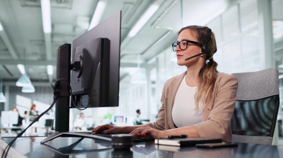 Vrouw met bril en headset werkt aan computer op kantoor met glazen wanden en bureaus op de achtergrond.