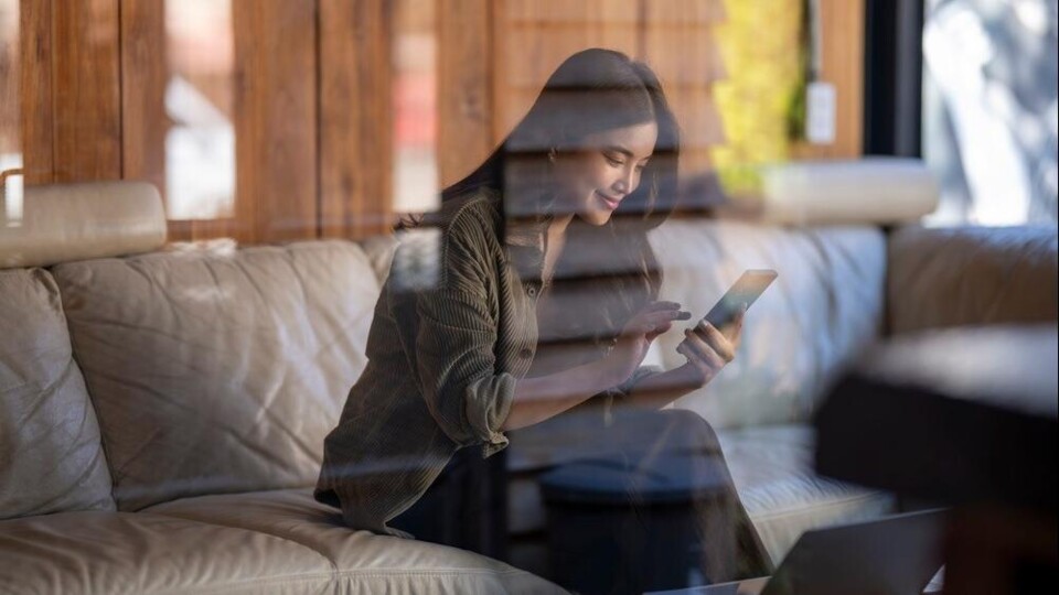 Vrouw zittend op een leren bank, gebruikmakend van een smartphone, met een laptop op tafel.