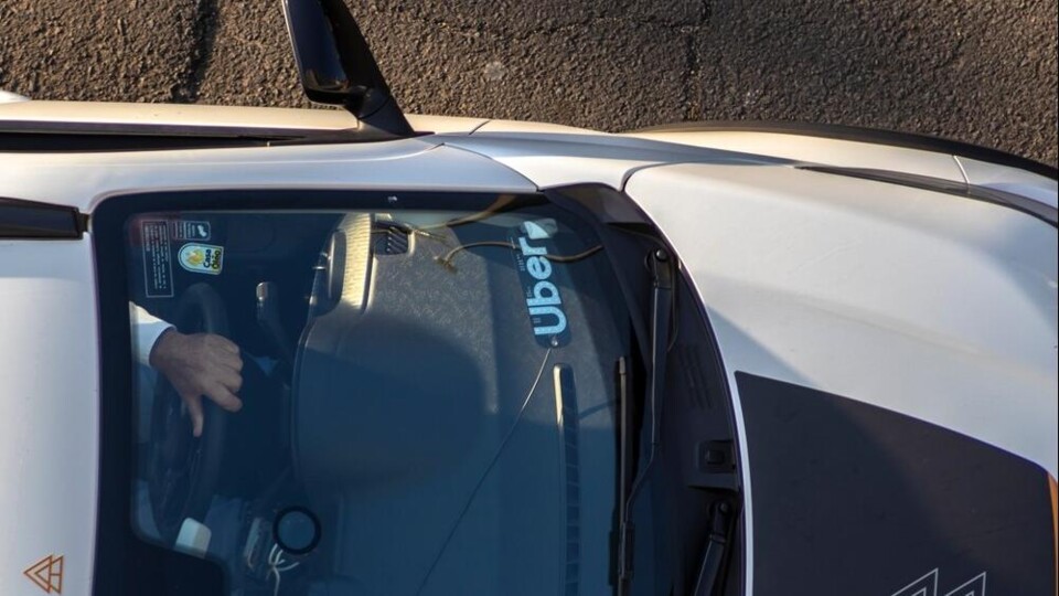 Witte auto met Uber-sticker op voorruit, bestuurder zichtbaar door raam, asfalt op achtergrond